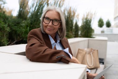 Gözlüklü, gri saçlı, kahverengi ceketli bir iş kadını sokakta terasta oturan bir iş ortağını bekliyor..