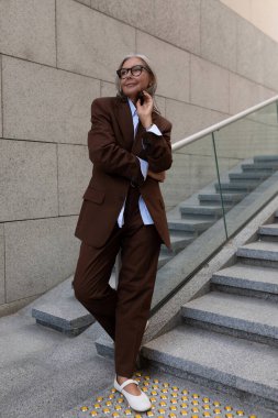 İnce gri saçlı olgun bir kadının portresi zarif kahverengi bir takım elbise giymiş bir binanın arka planında..