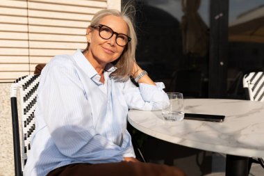 İş portresi. 60 yaşında, başarılı, beyaz saçlı, gözlüklü bir kadın. Terasta bir iş yemeğinde oturuyor..