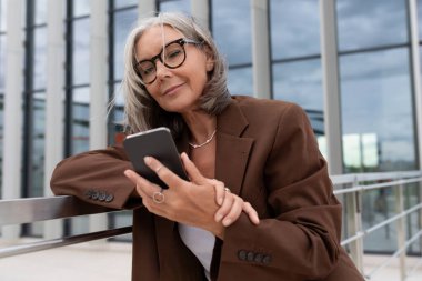 60 yaşında, narin, beyaz saçlı, şık giyinmiş bir iş kadını. Telefondaki mesajları kontrol ediyor..