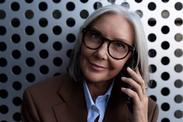 красивая седовласая успешная деловая женщина в очках в респектабельной одежде говорит по телефону на улице.