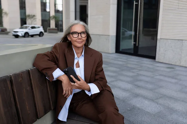 portrait of a confident successful 60 year old gray-haired business lady in glasses wearing a brown jacket on the street.