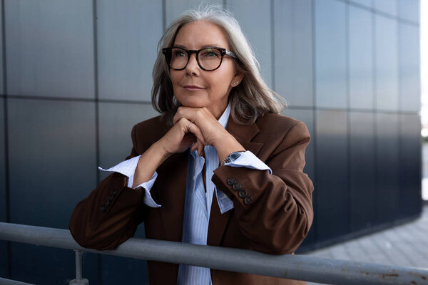 уверенная ухоженная старушка с седыми волосами в очках одета в коричневую куртку, а рубашка проводит обеденное время гуляя по улице.