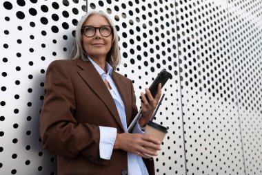 Başarılı, gri saçlı, şık giyimli büyük anne şehir caddesinde yürürken akıllı telefon kullanıyor..