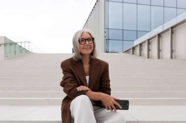 Gri saçlı, gözlüklü, şık ceketli mutlu yaşlı kadın şehirde dolanıyor ve şehirde merdivenlerde oturuyor..