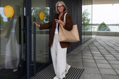 Gri saçlı ve gözlüklü mutlu yaşlı bayan şık giyinmiş bir şekilde iş merkezine girer..