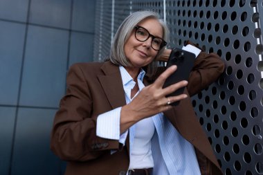 Kahverengi ceketli, beyaz saçlı, çekici bir iş kadını ve pantolonu sokakta iş konuşmak için akıllı telefon kullanıyor..