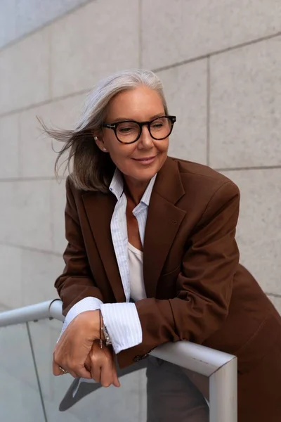 stock image successful free gray-haired business grandmother is dressed in a stylish brown suit against the backdrop of the stairs of the business center.