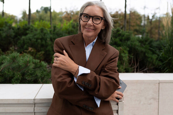 60-летняя успешная кавказская деловая леди одета в коричневый стильный костюм на фоне зелени города.