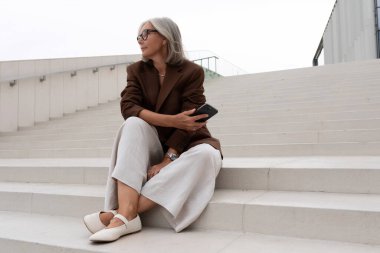 Kahverengi ceketli yaşlı, bakımlı, gri saçlı iş kadını ve pantolonu şehrin merdivenlerinde oturuyor..