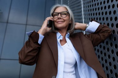 Kendine güvenen, saçları kısa saçlı, kahverengi ceketli, beyaz saçlı bir iş kadını ve telefonda konuşan bir pantolonu var..