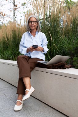 Gri saçlı, kahverengi takım elbiseli olgun bir iş kadını bankta oturmuş iş ortağını bekliyor..