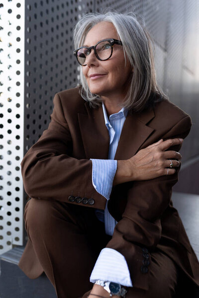 ухоженная пожилая седовласая деловая женщина в коричневой куртке и брюках ходит в городской среде.