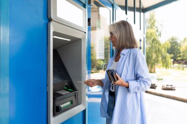 Olgun, gri saçlı bir kadın ATM 'de duruyor ve elektronik sistemi anlıyor..