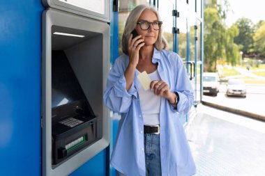 Yetişkin bir kadın ATM 'nin yanında dururken telefonla konuşuyor..