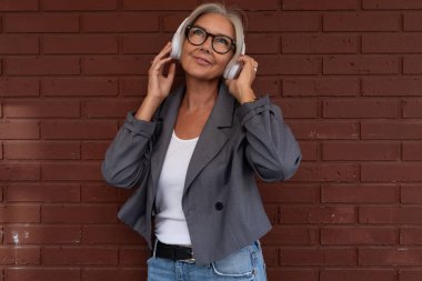 İnce, çekici, gözlüklü, gri ceketli ve kot pantolonlu bir kadın kulaklıkla müzik dinliyor..