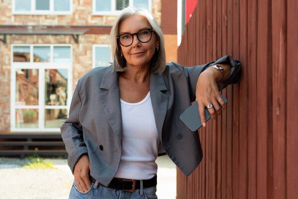 стильная зрелая женщина с седыми волосами и очками одета в серый пиджак прогуливаясь по городу.