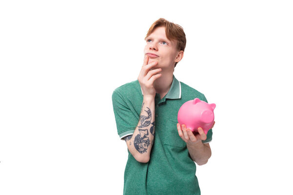 joyful happy young red-haired guy in a green t-shirt holds a piggy bank with money and thinks where to spend.