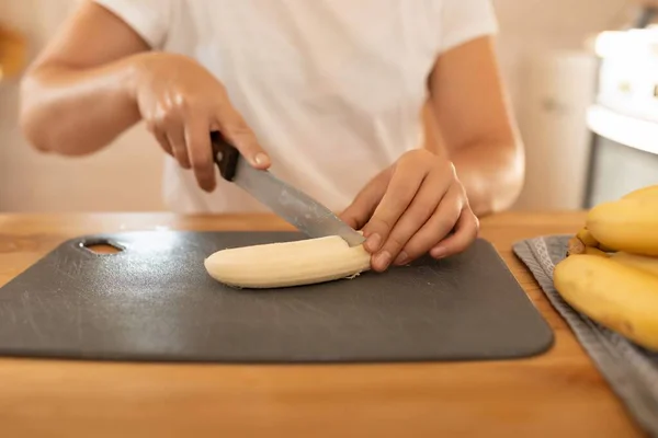 a woman slices a banana with a knife on a board in the kitchen.