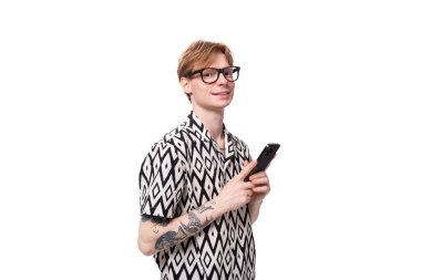 Gömlek giymiş kızıl saçlı genç beyaz bir adam elinde bir telefon tutuyor..