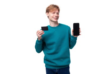 Mavi kazaklı, kızıl saçlı beyaz bir çocuk elinde bir akıllı telefon ve bir kredi kartı tutuyor..