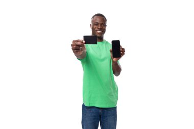 Açık yeşil tişörtlü ve kot pantolonlu genç, bakımlı Afrikalı bir adam bir kredi kartı ve bir akıllı telefon gösteriyor..