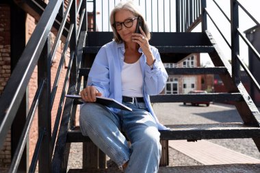 Gri saçlı güzel, olgun bir kadın dizüstü bilgisayarla sokakta otururken bir telefon görüşmesi yapıyor..