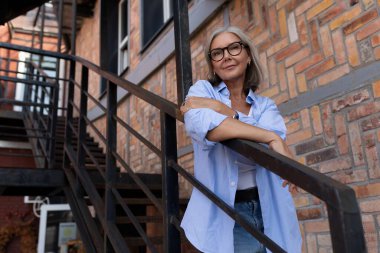 Yazlık kıyafetli, olgun, kır saçlı bir kadın şehir caddesinde yürüyor..