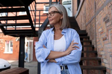 Kendine güvenen, gri saçlı yaşlı bir kadın şık bir şekilde şehirde dolaşıyor ve merdivenlerde duruyor..