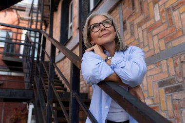 Yazlık kıyafetli, olgun, kır saçlı bir kadın şehir caddesinde yürüyor..