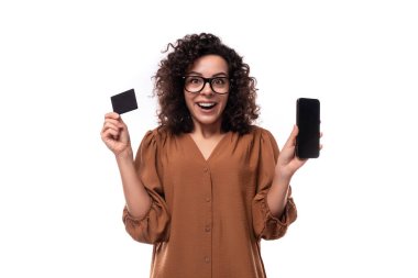 Genç, neşeli, beyaz, kıvırcık, kahverengi tişört giymiş esmer kadın elinde plastik bir kart ve akıllı bir telefon tutuyor..
