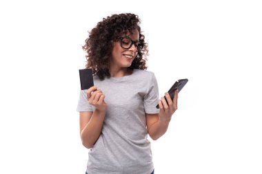 Hoş, ince kıvırcık saçlı, siyah saçlı, sıradan gri tişörtlü, elinde akıllı telefon ve plastik kredi kartı olan bir kadın..
