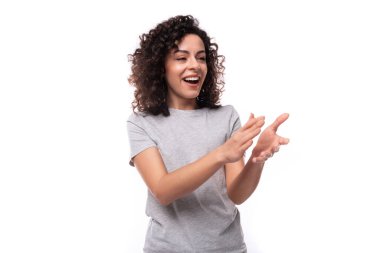 Beyaz arka planda gri tişörtlü, kıvırcık saçlı, hoş, genç, Avrupalı esmer bir kadının portresi..