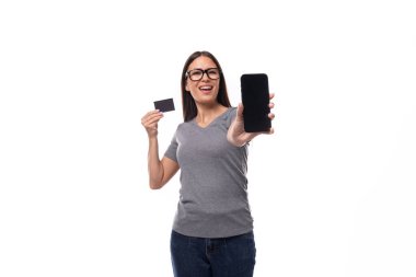 Gri tişört giymiş genç esmer Avrupalı kadın beyaz arka planda bir akıllı telefon ve plastik kart ekranı gösteriyor..