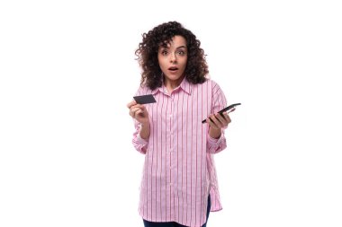Genç, otantik, kıvırcık, esmer, pembe çizgili gömlek giymiş kadın kredi kartı ve akıllı telefon tutarken şaşırdı..