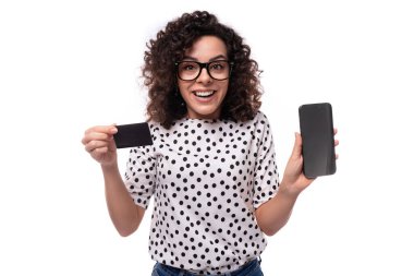 Siyah kıvırcık saçlı, puantiyeli bluzlu genç, bakımlı bir bayan elinde plastik bir kart ve akıllı bir telefon tutuyor..