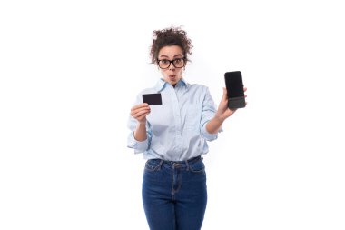 Beyaz arka planda plastik kredi kartı ve akıllı telefon tutan kıvırcık saçlı, güler yüzlü beyaz kadın..