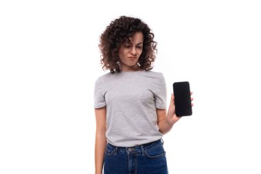 Genç üzgün, esmer, kıvırcık, gri tişörtlü kadın. Beyaz arka planda cep telefonu ekranını gösteriyor..