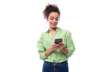 Atkuyruklu kıvırcık saçlı çekici, genç, kıvırcık saçlı kadın yeşil gömlek ve kot pantolon giymiş telefonda mesaj yazıyor..