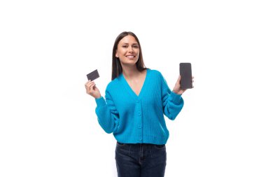 Mavi hırka giymiş, elinde banka kartı modeli ve beyaz arka planda fotokopi alanı olan akıllı telefon tutan genç, pozitif esmer bir kadın..