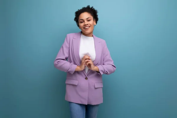 young cute hispanic business woman with curly hair in a lilac jacket smiling on a blue background with copy space.