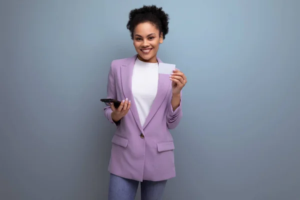 young successful latin woman with afro hair dressed in a lilac jacket shows a mock-up of a bank card and a smartphone.