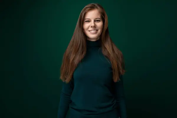 Cute modest woman with brown natural hair posing shyly on dark green background with copy space.