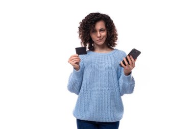 Mavi kazak giymiş güzel kıvırcık esmer kadın elinde plastik bir banka kartı ve akıllı telefon tutuyor..