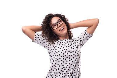 Kıvırcık saçlı genç bir kadın beyaz arka planda benekli bir yaz bluzu giyiyor..