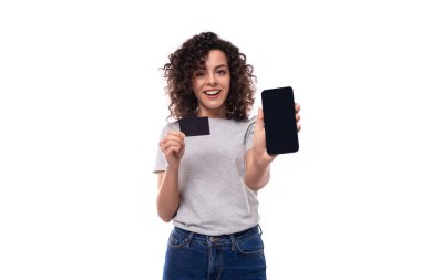 Gri, esmer, kıvırcık saçlı, gri tişörtlü genç bir kadın, arka planda fotokopi alanı olan plastik bir kart ve akıllı bir telefonu gösteriyor..