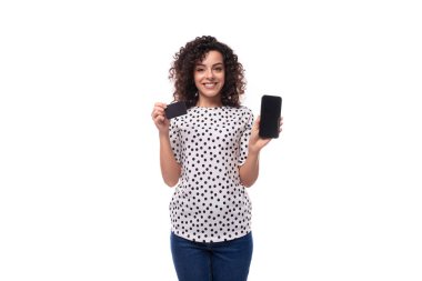 young dark-haired caucasian woman dressed in a blouse demonstrates a plastic credit card and a smartphone.