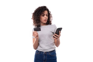 young charming brunette curly lady in a gray basic t-shirt holding a mockup of a plastic card and a smartphone on a white background with copy space.