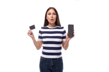 charming young european woman with black straight hair in a striped black and white t-shirt and jeans holds a credit card with a mockup and a smartphone with a blank screen.