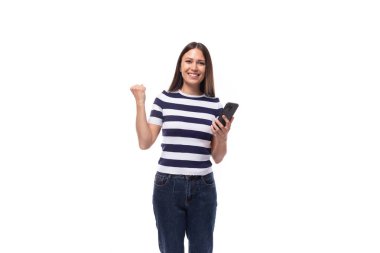 young brunette woman model dressed in a striped t-shirt and jeans rejoices holding a phone in her hand.